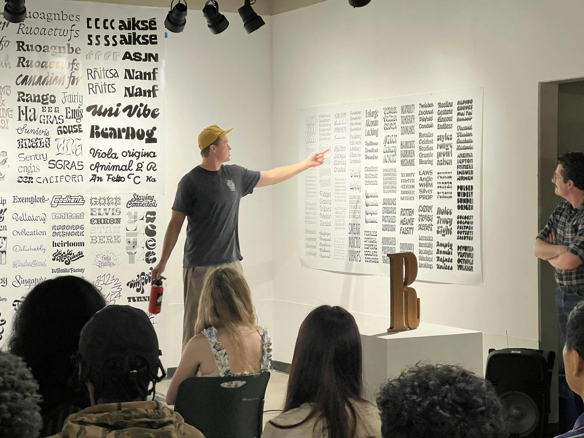 Audience members look in James Edmondson's direction as he points to typefaces on papers covering a wall.