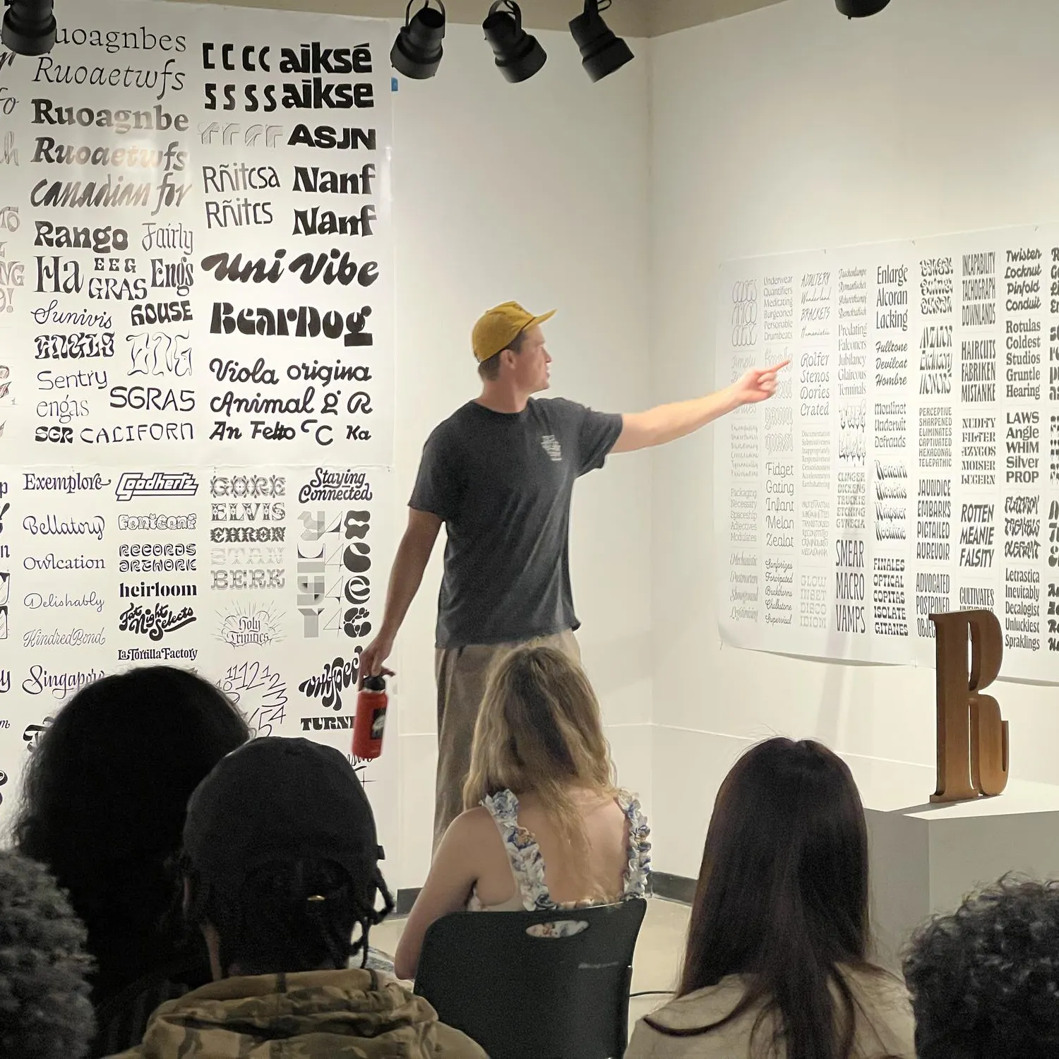 Audience members look in James Edmondson's direction as he points to typefaces on papers covering a wall.
