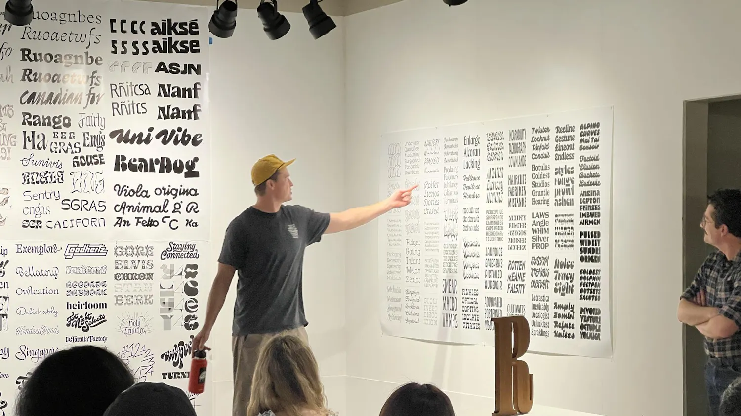 Audience members look in James Edmondson's direction as he points to typefaces on papers covering a wall.