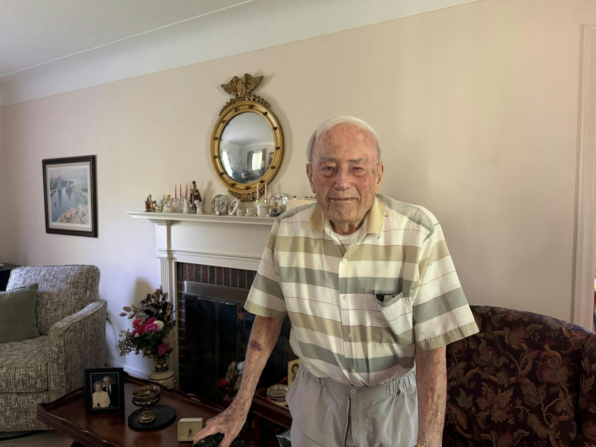 A man stands in his living room in front of a fireplace, holding on to his walker. 