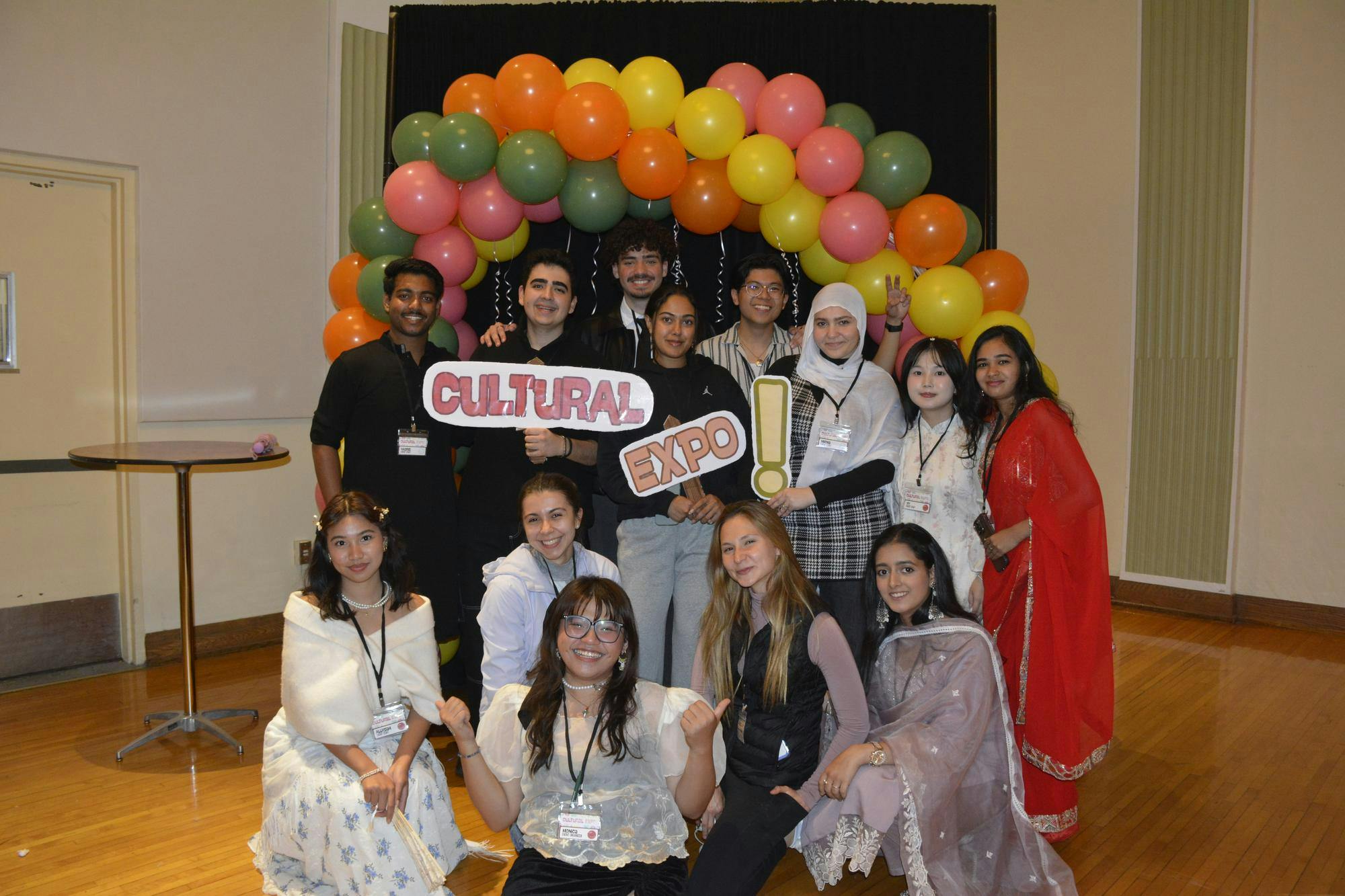 A group of students stand and kneel in front of a colorful balloon arch and some students hold signs that spell out "Cultural Expo!"