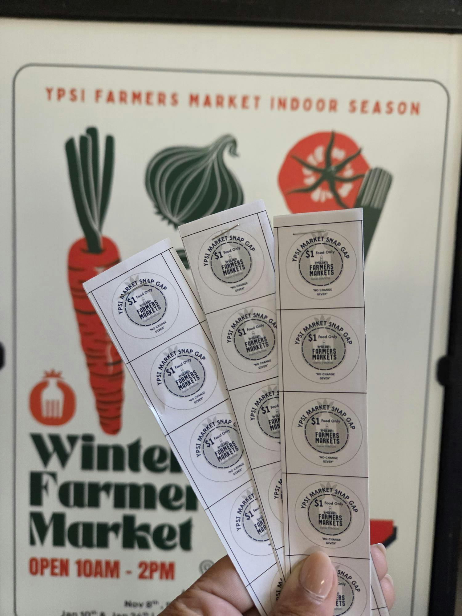 Three strips of white paper containing SNAP Gap tokens. Each token is in the shape of a circle. Blue letters at the top spell out "Ypsi Market Snap Gap," followed by "$1 Food Only" on the next line. "Farmers Markets" is spelled out on the line underneath, and at the bottom, blue letters spell out "No change given."