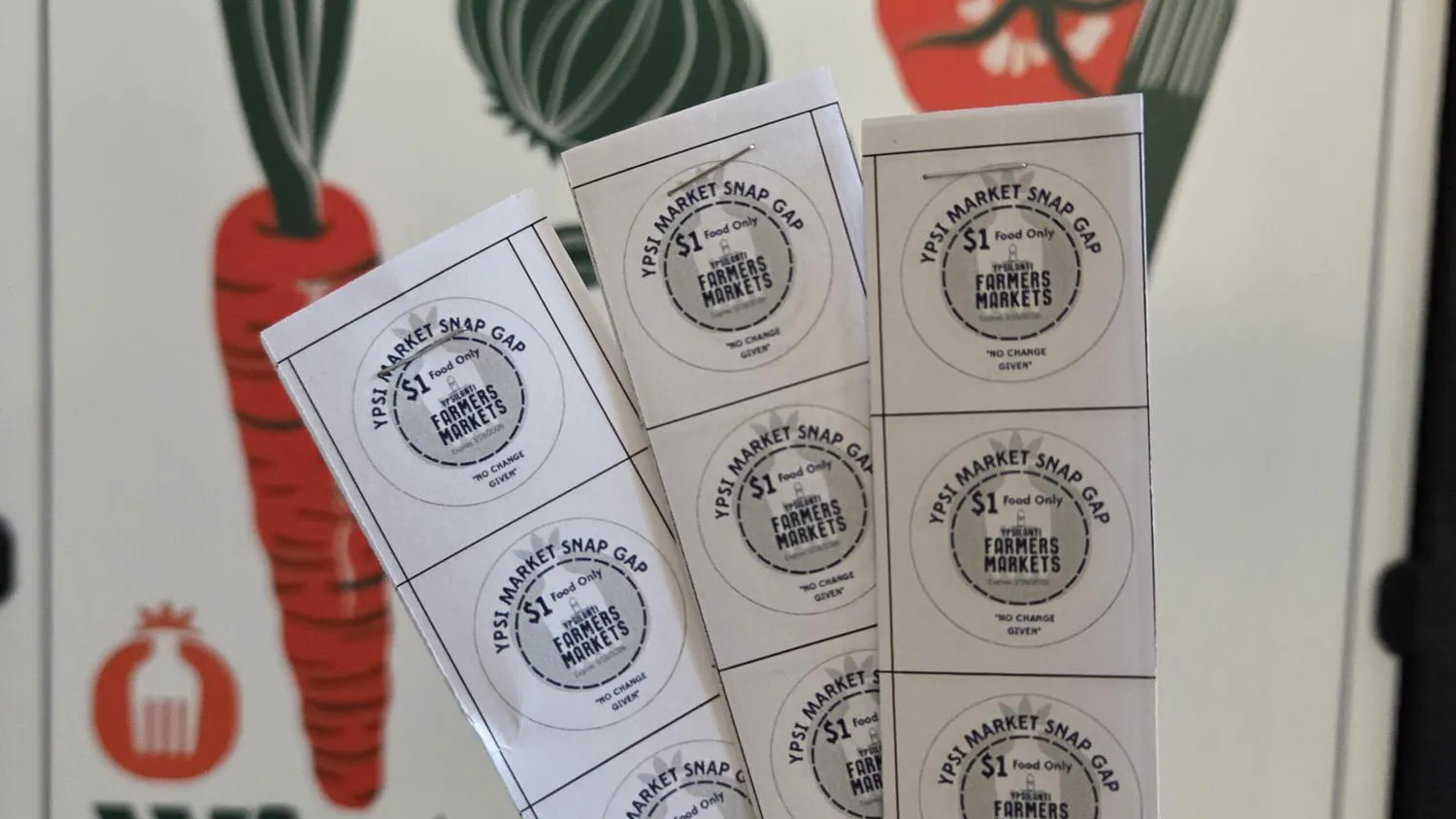 Three strips of white paper containing SNAP Gap tokens. Each token is in the shape of a circle. Blue letters at the top spell out "Ypsi Market Snap Gap," followed by "$1 Food Only" on the next line. "Farmers Markets" is spelled out on the line underneath, and at the bottom, blue letters spell out "No change given."