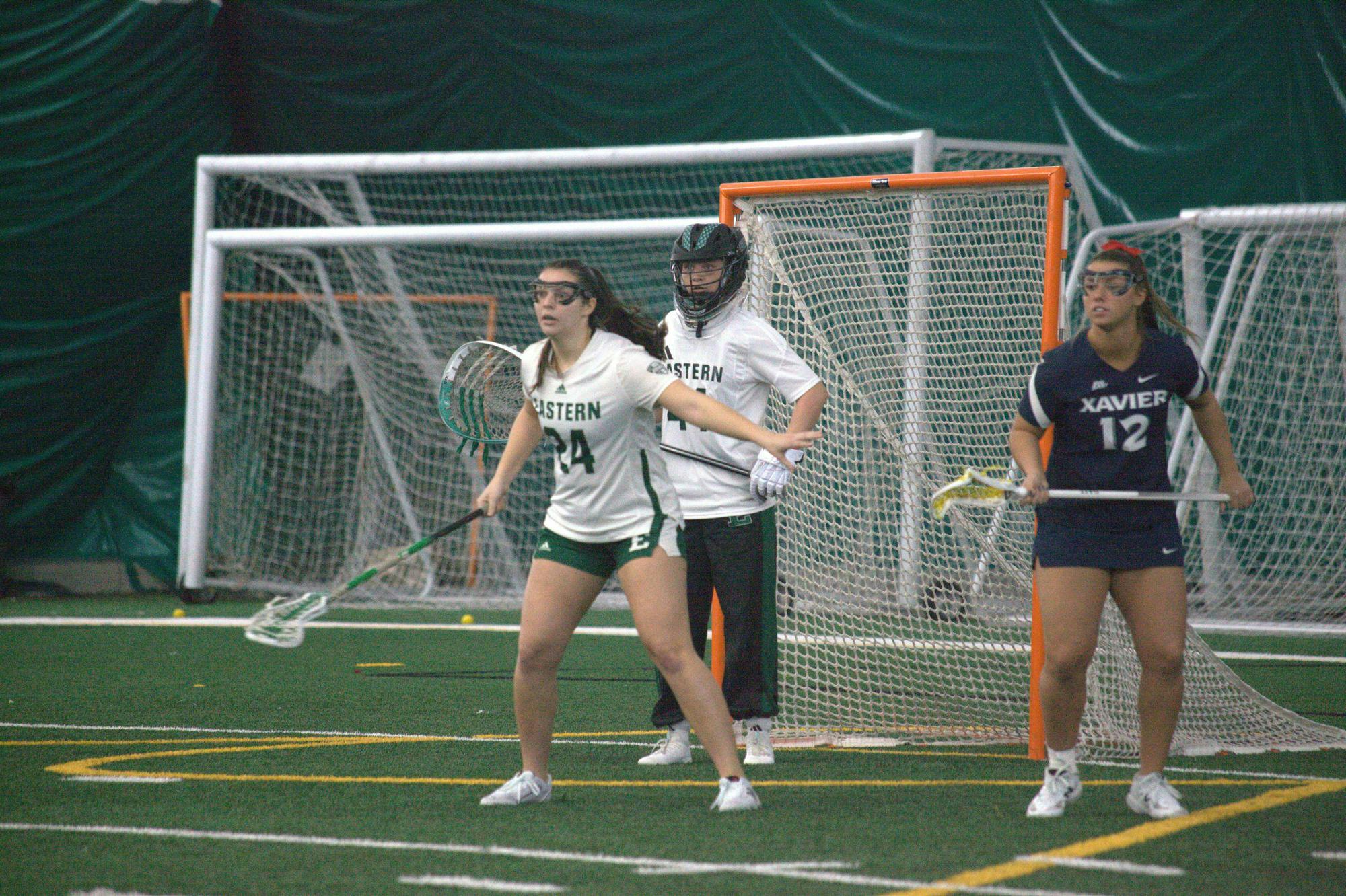 Two women's lacrosse players guard goal while a member of the opposing team stands off to the side.
