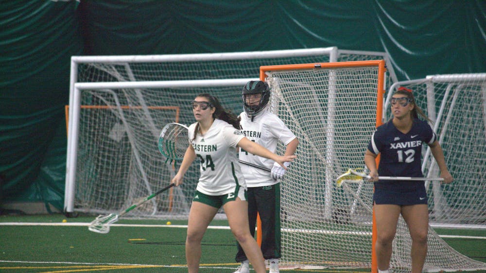 Two women's lacrosse players guard goal while a member of the opposing team stands off to the side.