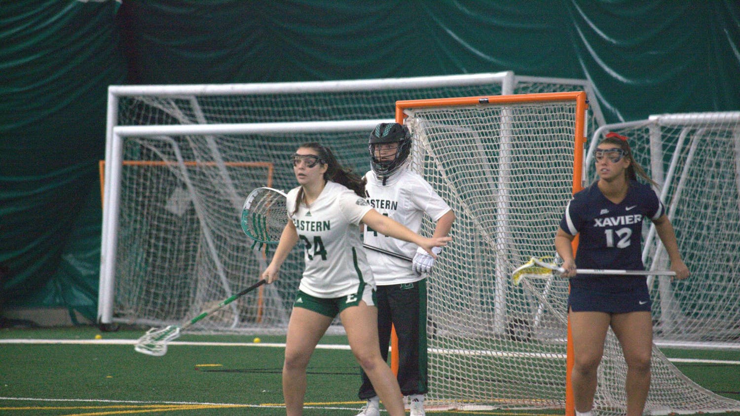 Two women's lacrosse players guard goal while a member of the opposing team stands off to the side.