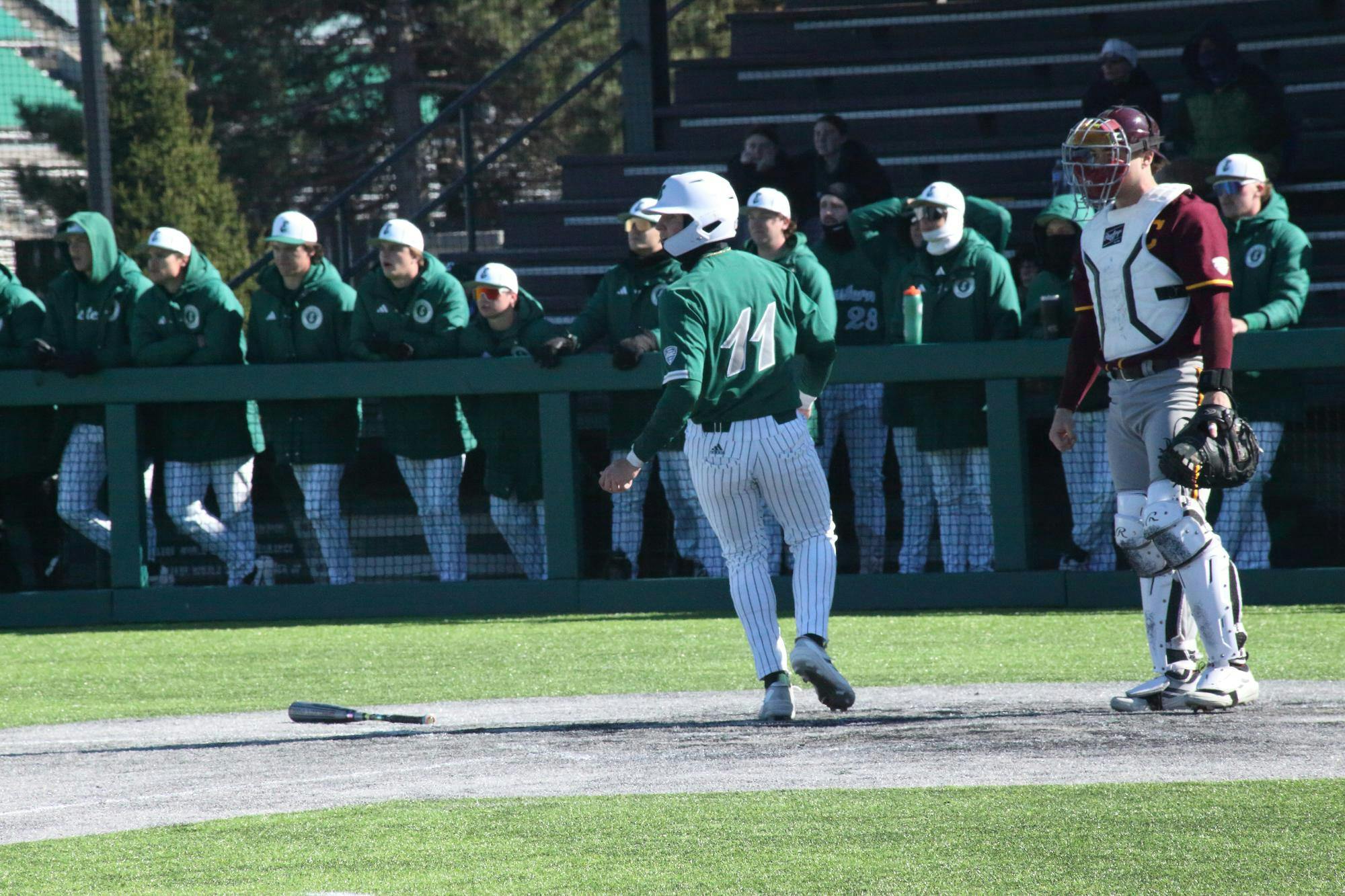 Baseball player mid-jog at home base at outdoor game. A large group of teammates stand in the background, watching another player who is out of frame.