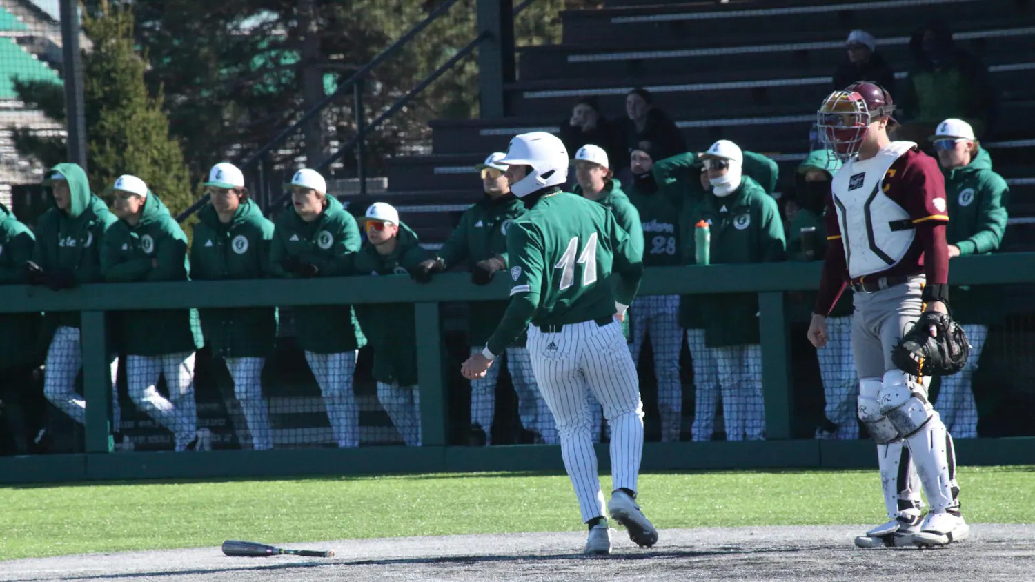 Baseball player mid-jog at home base at outdoor game. A large group of teammates stand in the background, watching another player who is out of frame.