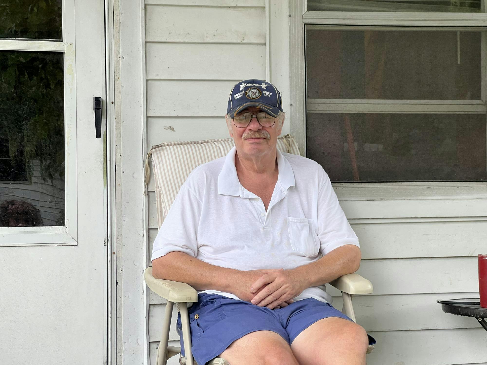A man sits on a cream-colored chair on the porch of a white house.