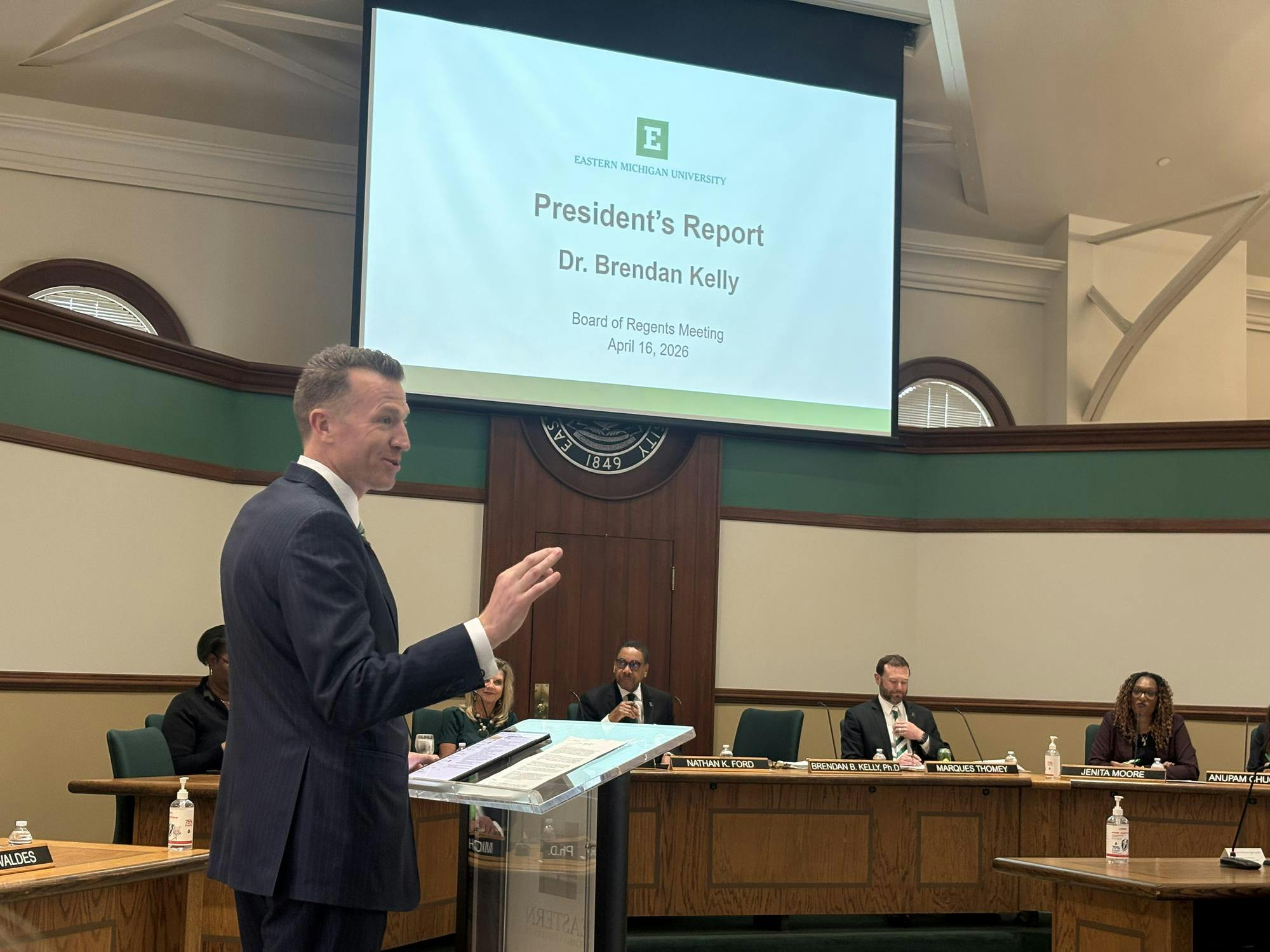 Brendan Kelly speaks from behind a clear, transparent plastic podium as Board of Regents members listen from a long, shared desk in the background.