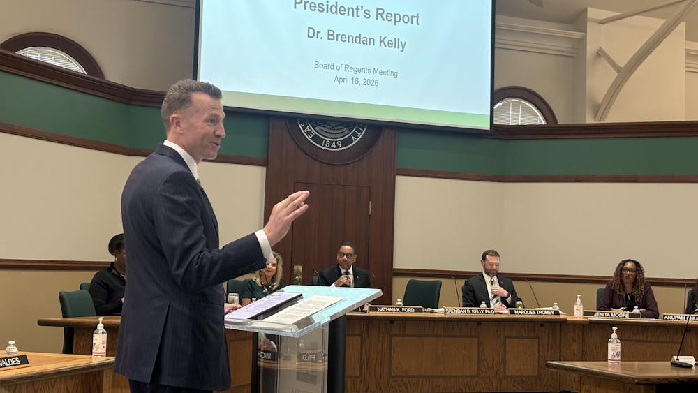 Brendan Kelly speaks from behind a clear, transparent plastic podium as Board of Regents members listen from a long, shared desk in the background.