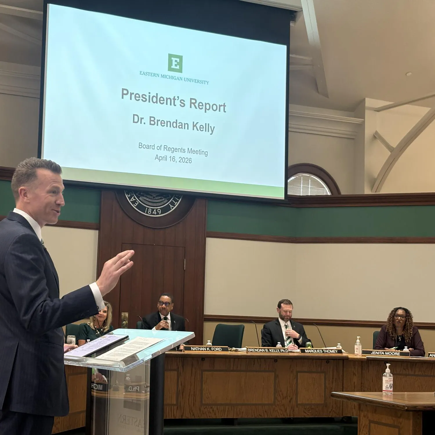 Brendan Kelly speaks from behind a clear, transparent plastic podium as Board of Regents members listen from a long, shared desk in the background.
