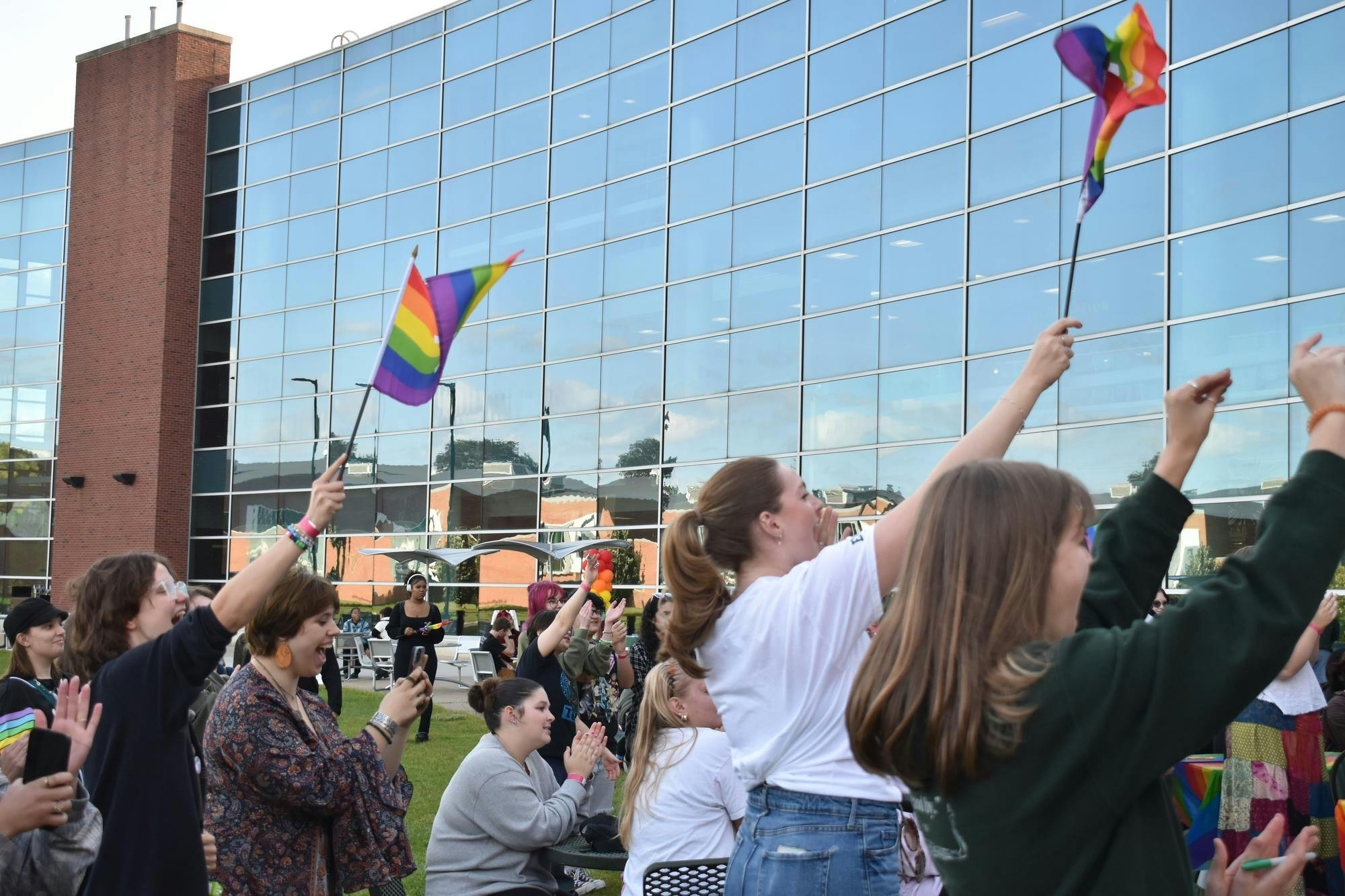 Multiple students are stood around the Student Center patio cheering and waving pride flags in the air while smiling. 