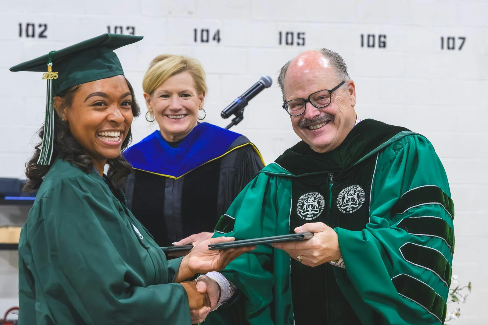 Wearing green robes, EMU's president James Smith passes a diploma to student Andrea Bracy while a third person watches from behind. Everyone is smiling.