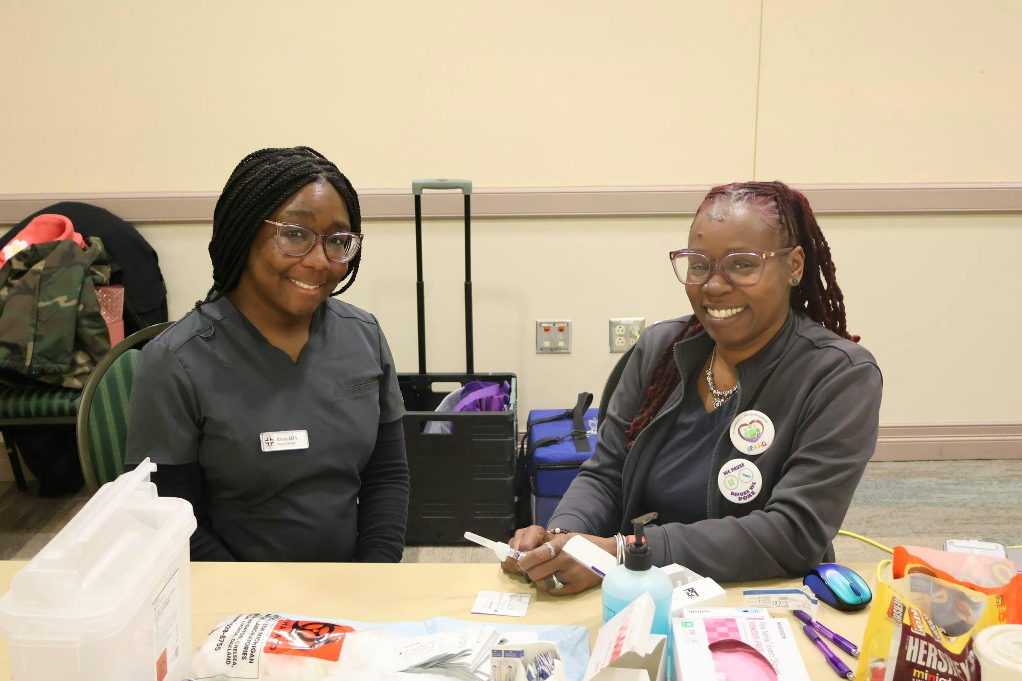 Two women in scrubs smiling behind a table with medical supplies.