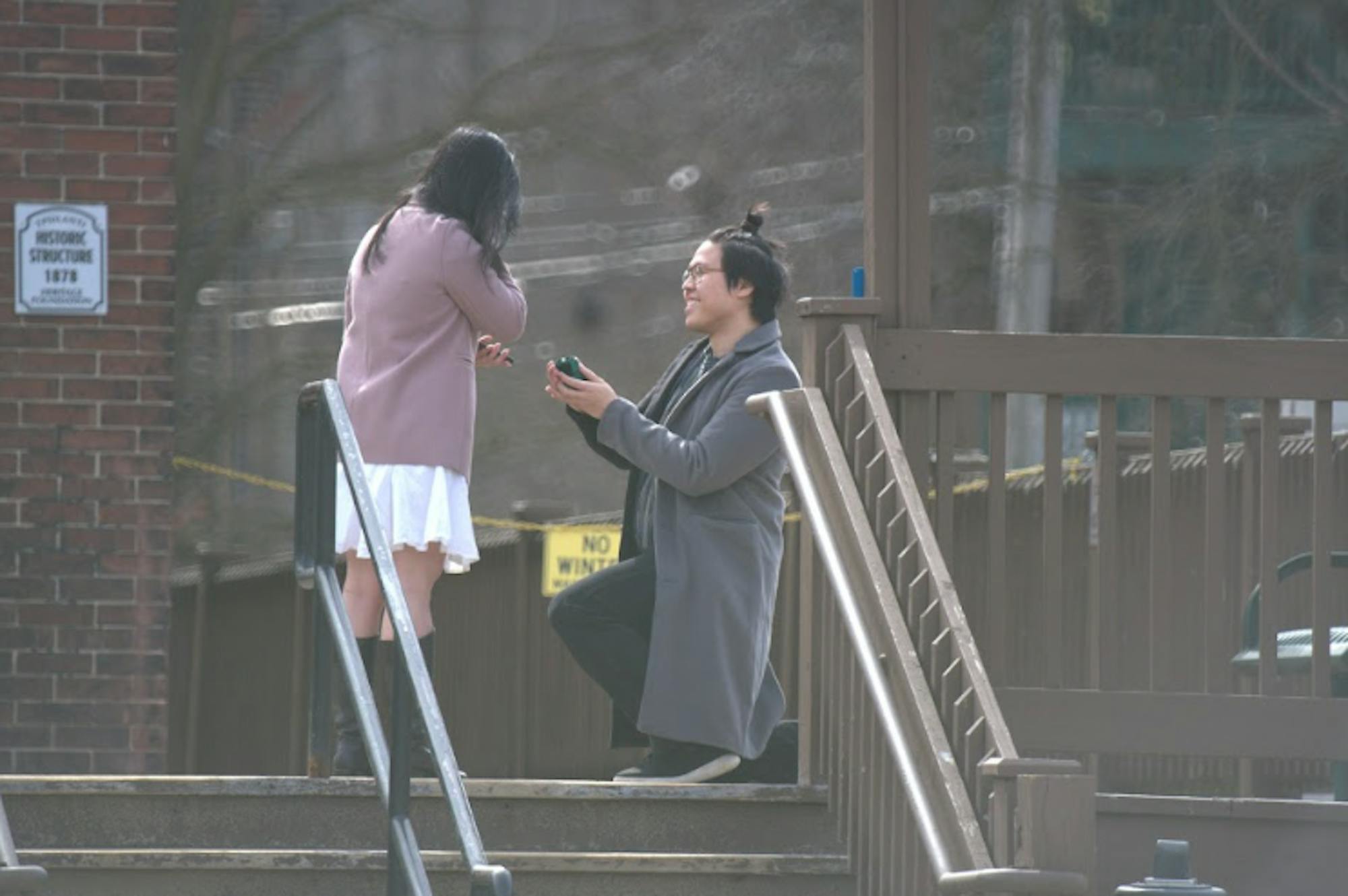On the right, Christopher Kok is on one knee, holding up a ring box and proposing to Julian Ho who stands in front of him. 