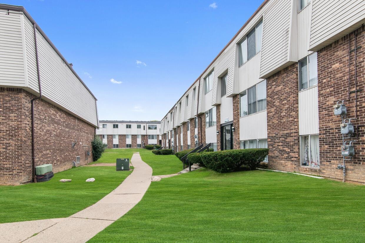 This picture shows the Arbor One apartment buildings in Ypsilanti, Michigan. The bottom part of the buildings is made from bricks in different shades of red and orange, while the upper part features wood siding. A sidewalk runs through the middle of the picture, and there are green bushes and grass around the buildings.