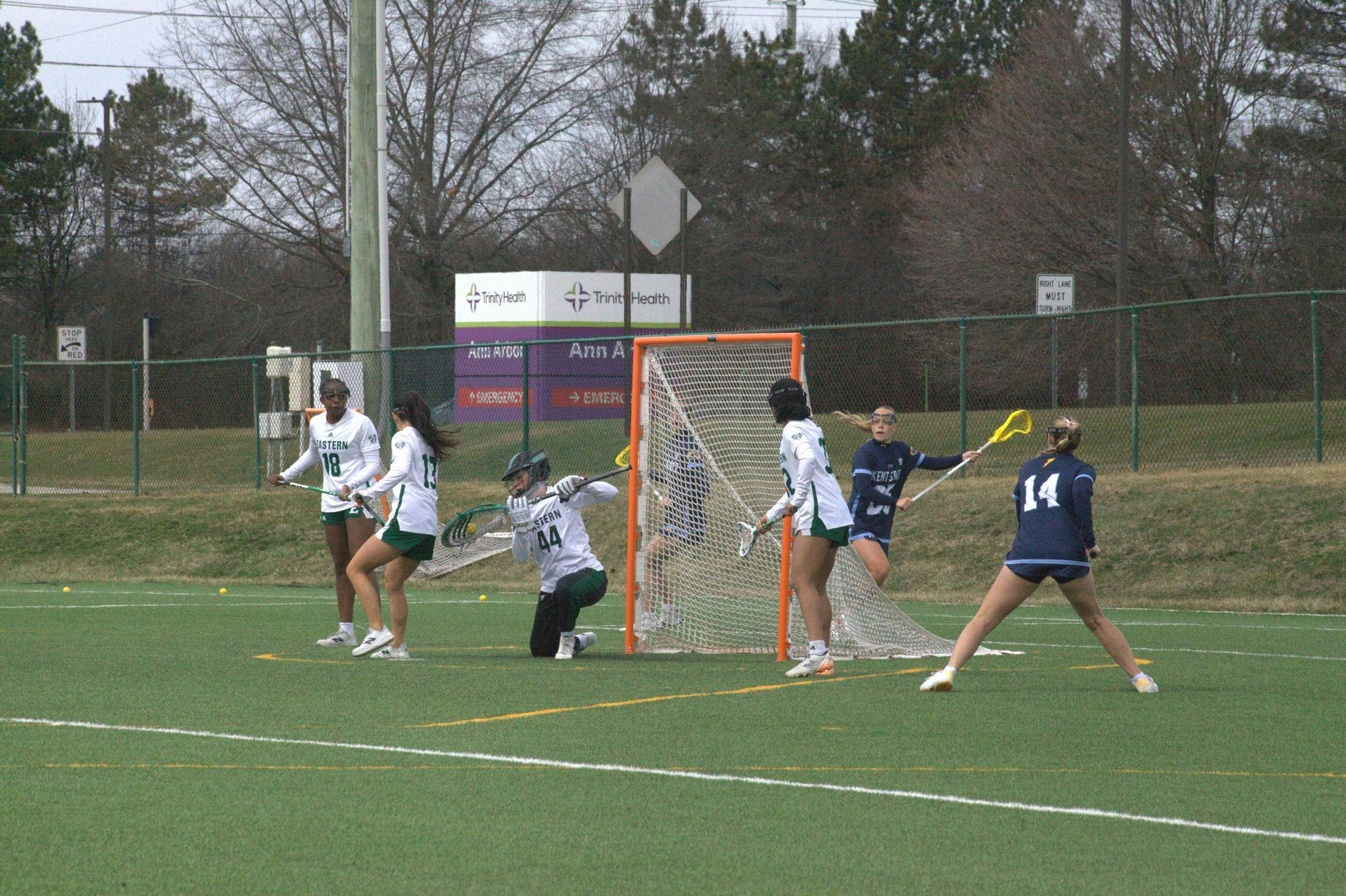 A women's lacrosse goalie intercepts a score from the opposing team on an outdoor lacrosse field with defensive teammates standing around the goal.