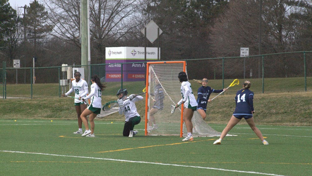 A women's lacrosse goalie intercepts a score from the opposing team on an outdoor lacrosse field with defensive teammates standing around the goal.