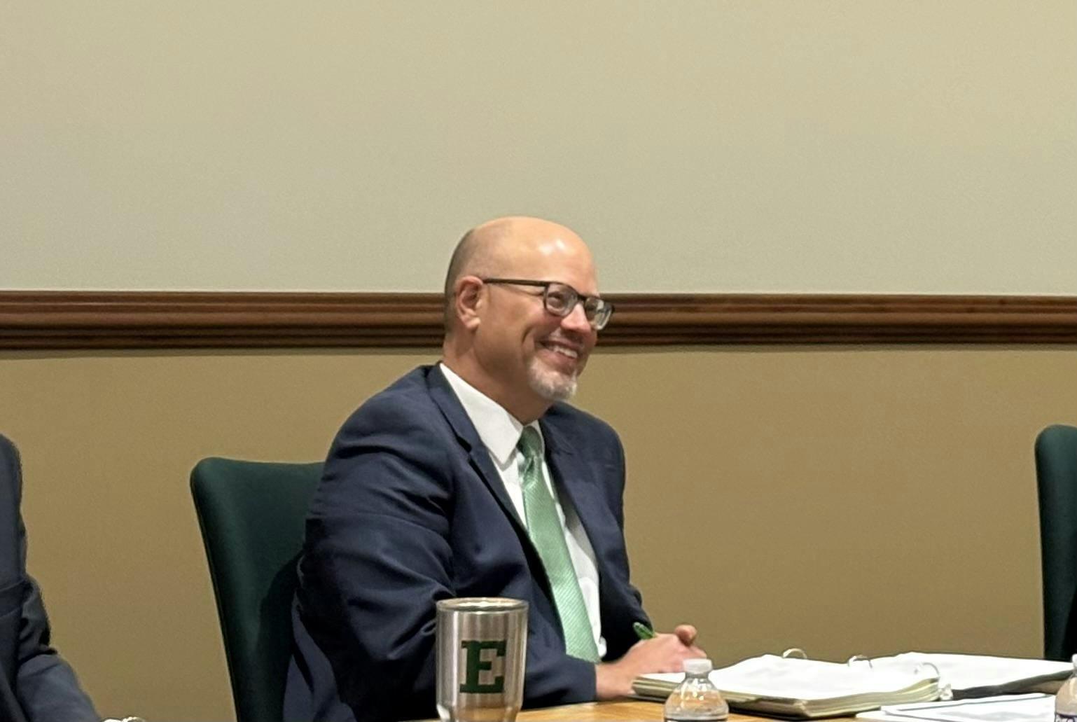  David Schecter sits at a wooden desk during a meeting, smiling.