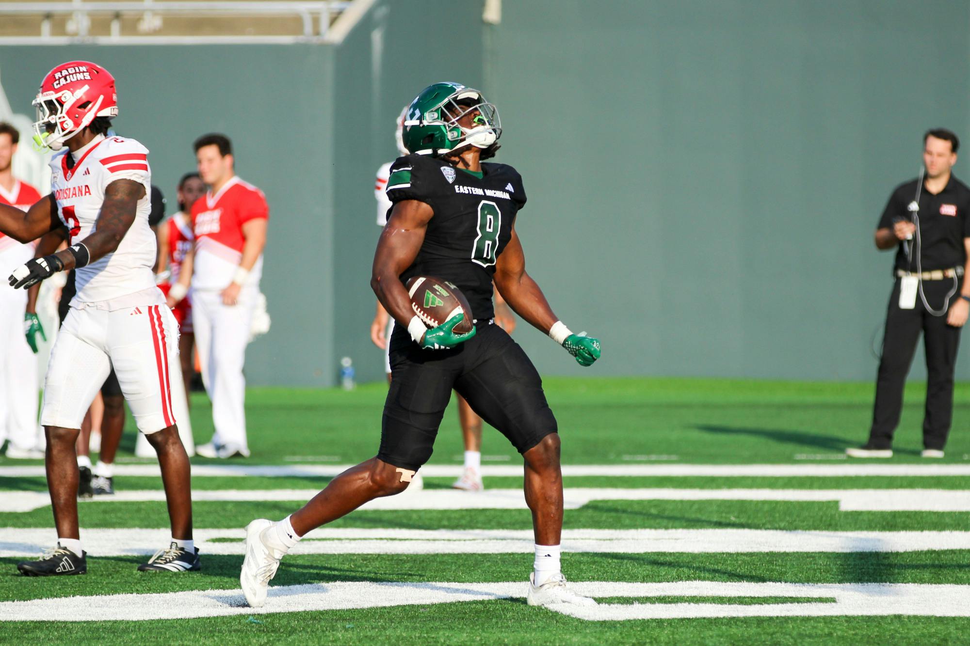 An Eastern Michigan ball carrier rejoices in the end zone after scoring a touchdown, dressed in Eastern Michigan's black-out uniform and holding the ball in his hand. 
