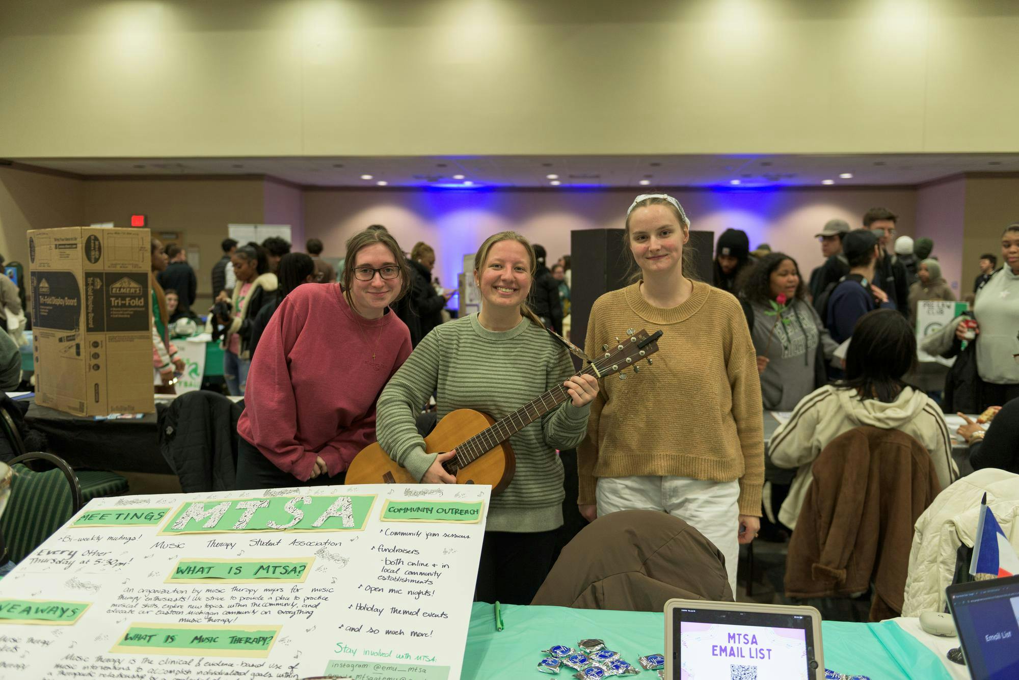 Halie holds a guitar in the middle while Amanda and Katelyn lean in for the picture with her while standing behind their booth with a green information poster for their club.