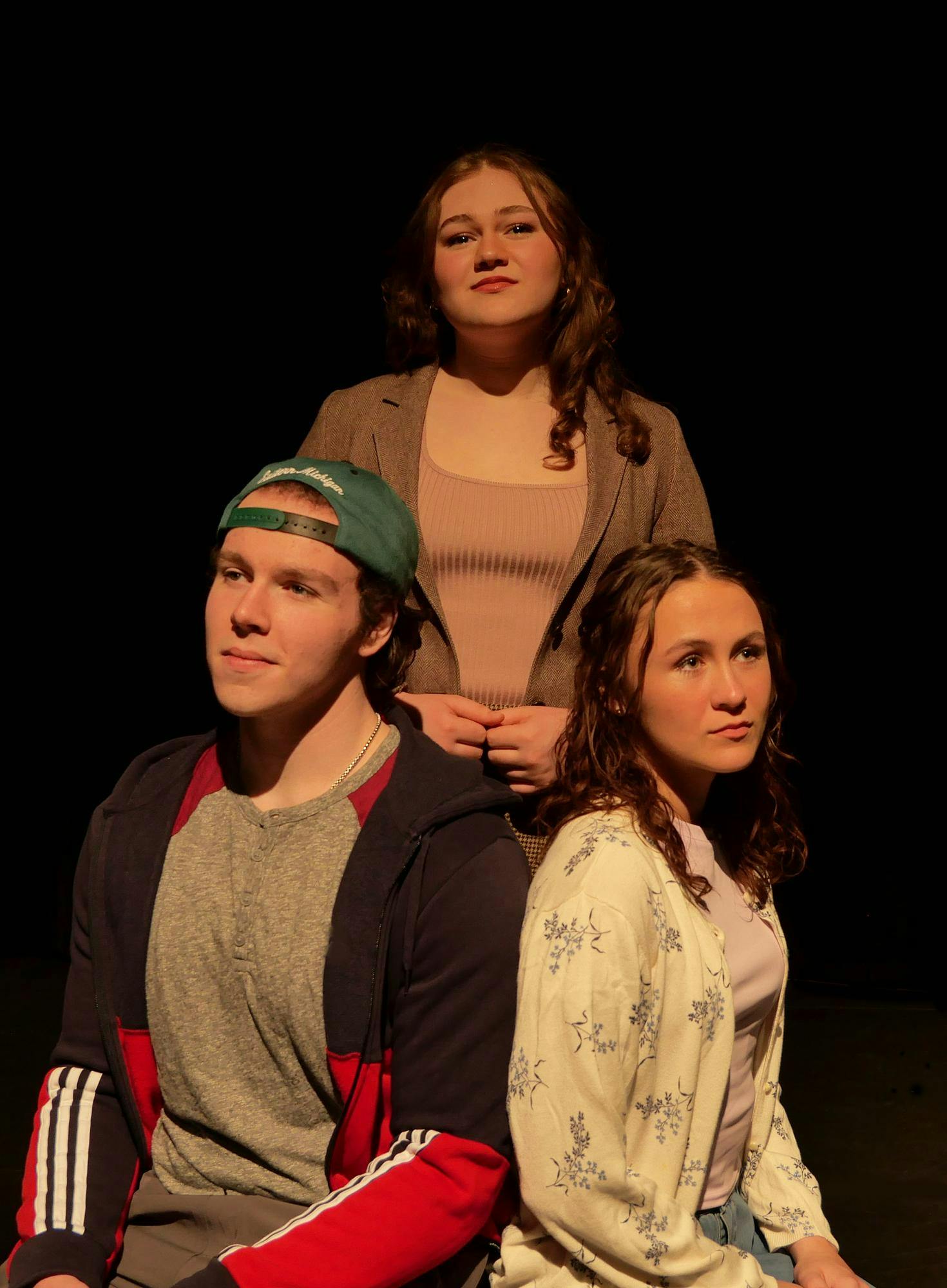 Three people are positioned together, with two sitting in front and one standing behind the others. The stage lights softly illuminate their faces as they all stare off into the distance.