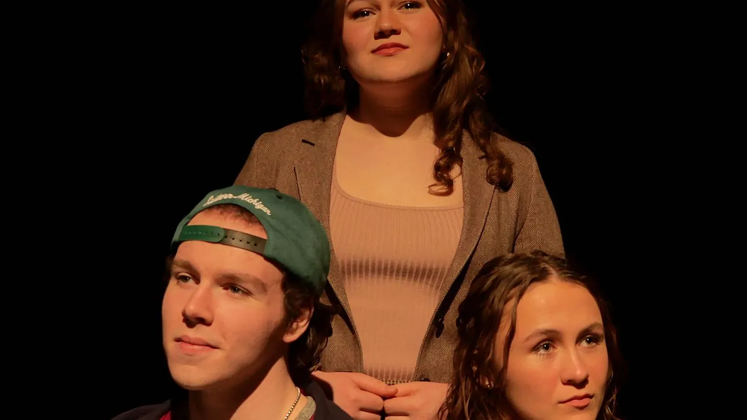 Three people are positioned together, with two sitting in front and one standing behind the others. The stage lights softly illuminate their faces as they all stare off into the distance.