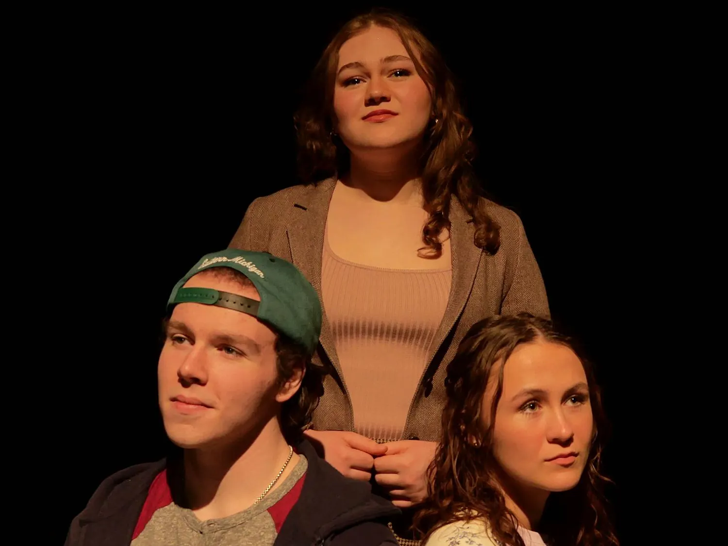 Three people are positioned together, with two sitting in front and one standing behind the others. The stage lights softly illuminate their faces as they all stare off into the distance.