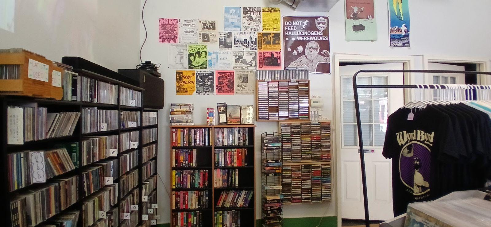 Inside of a store that has shelves of books to the right, movies and CDs in the center, and t-shirts on the left.