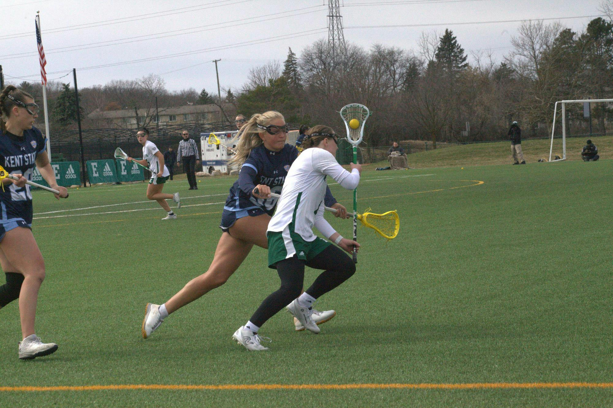 Women's lacrosse players of different teams run on the field near while one tries to knock the ball out of the other's stick.