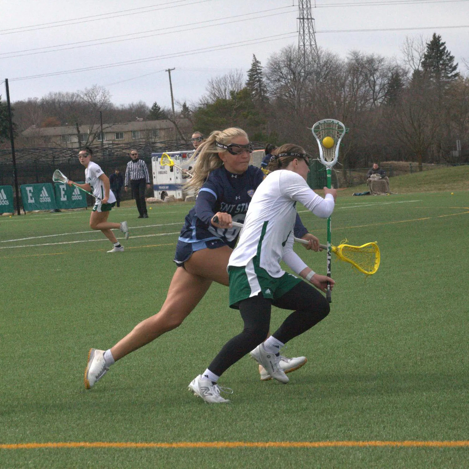 Women's lacrosse players of different teams run on the field near while one tries to knock the ball out of the other's stick.