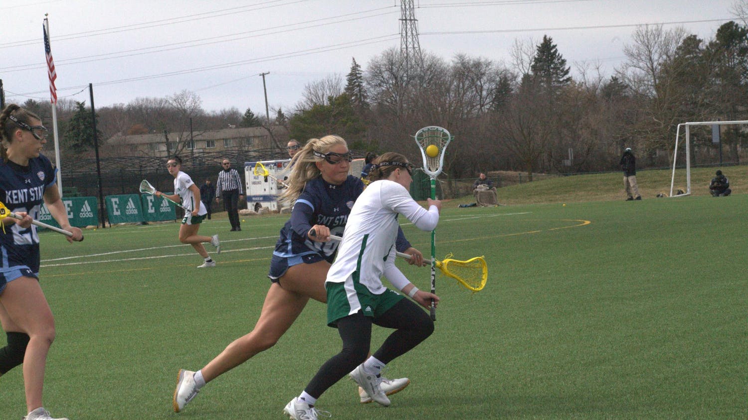 Women's lacrosse players of different teams run on the field near while one tries to knock the ball out of the other's stick.