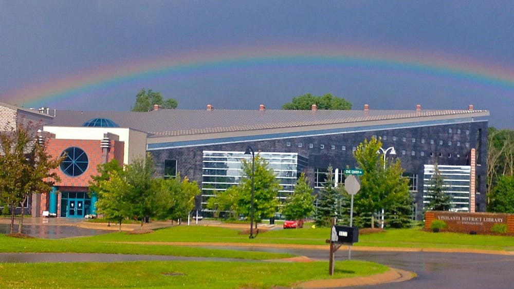 A building with a red-and-blue-brick entrance. Extending from the right side of the entrance is a long gray-colored section of the building. Several green trees frame the entrance to the building, and there is green grass in the foreground of the photo. Overhead, the sky is blue, and there is a rainbow visible over the building.