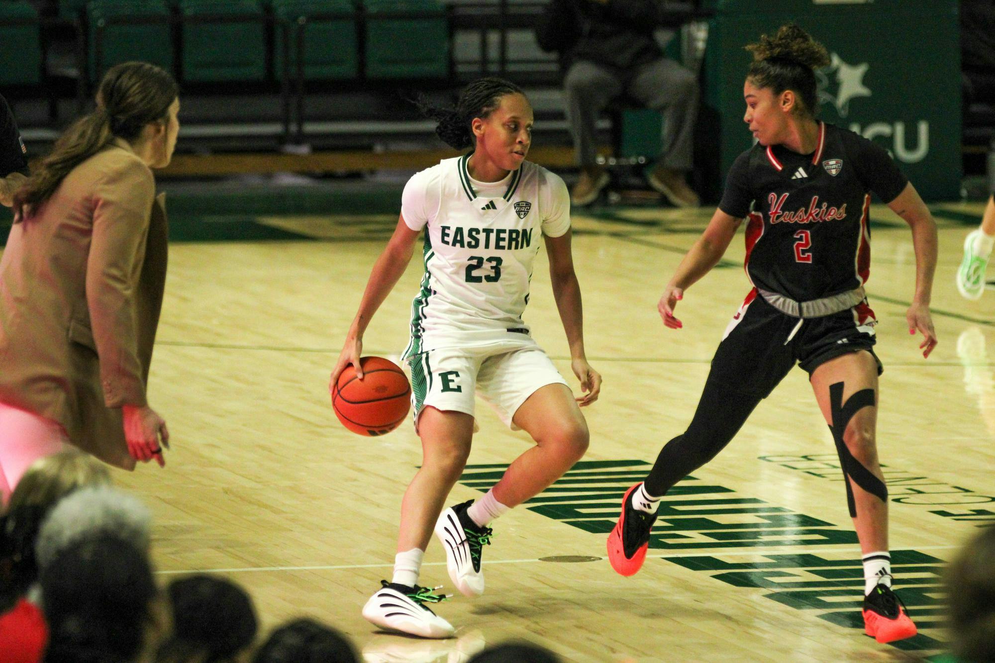 EMU player #23, in a green jersey, dribbling the basketball with pressure from NIU player, #2. 