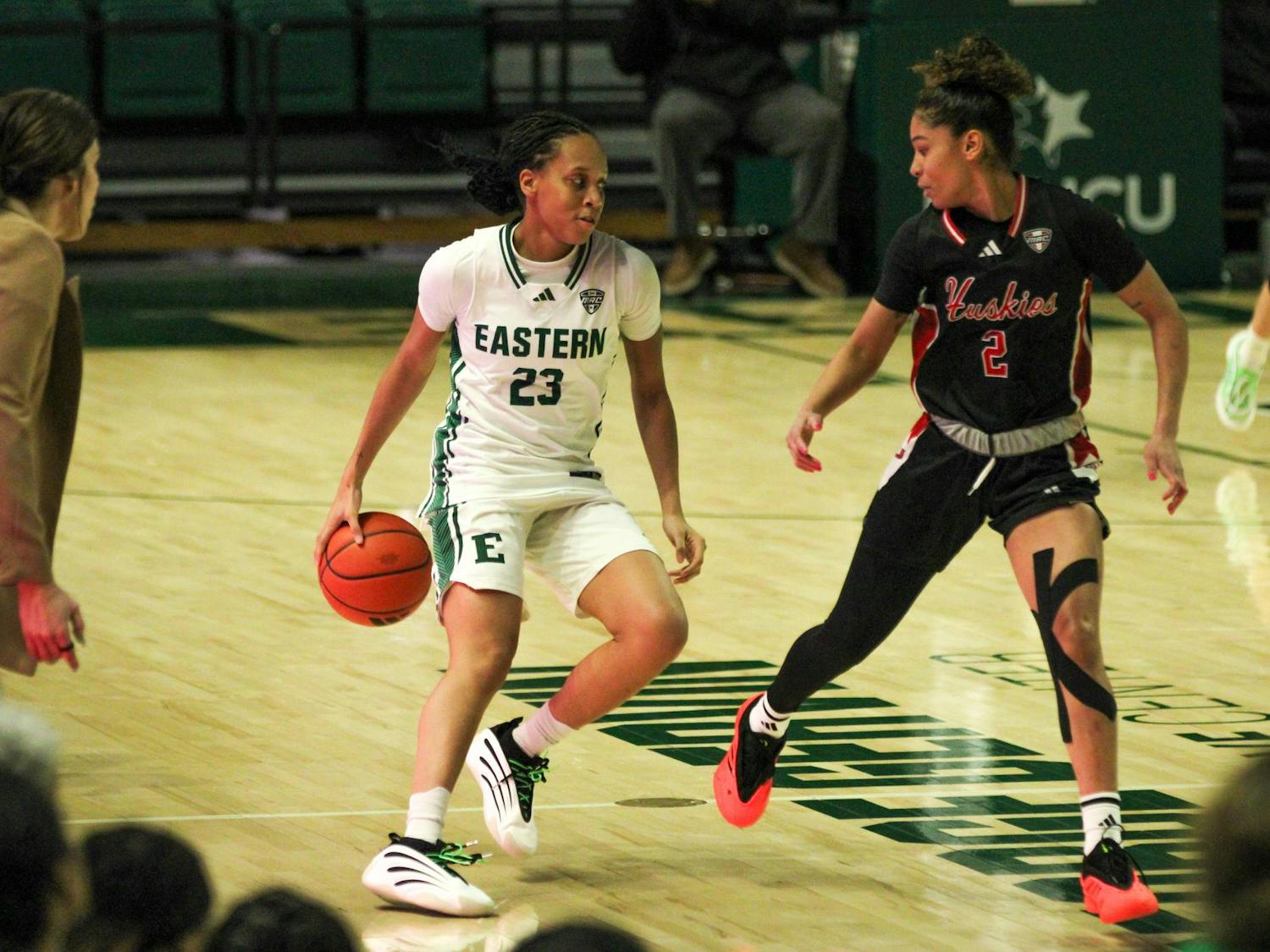 EMU player #23, in a green jersey, dribbling the basketball with pressure from NIU player, #2.