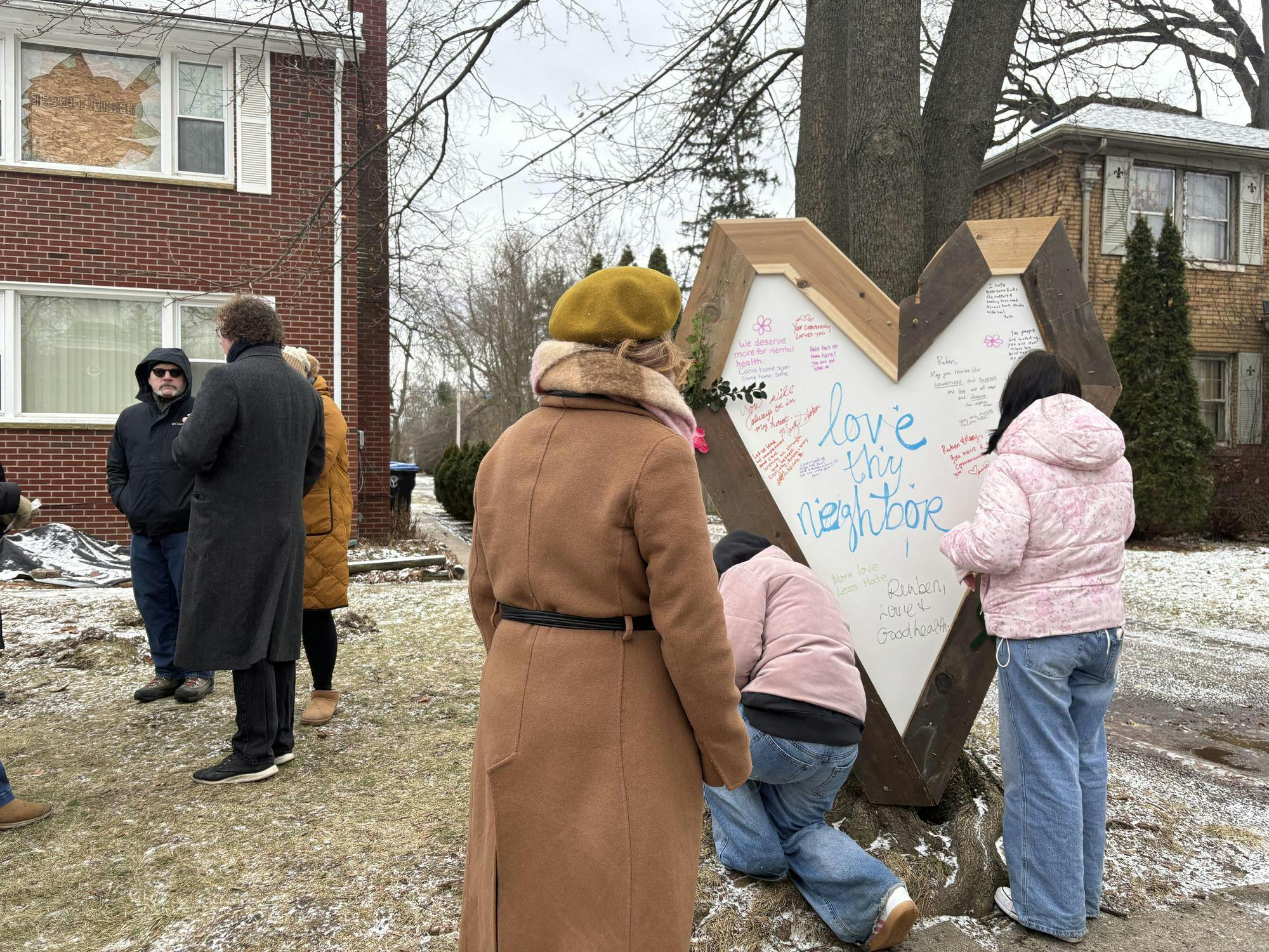 A large, hand-crafted heart-shaped sign with "Love thy neighbor" written in the center, along with other messages of support, stands in the yard of a damaged and condemned two-story brick house where several community members gather to add messages to the sign.