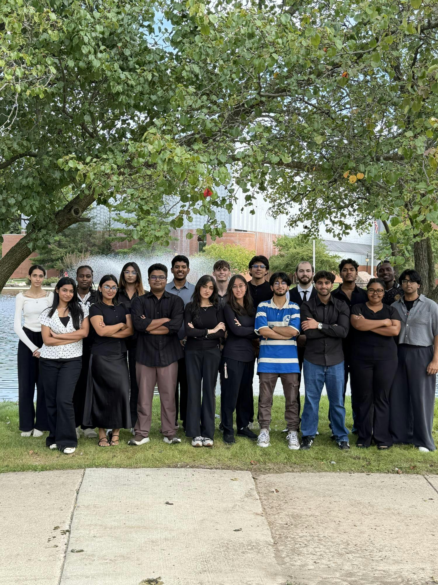 A group of 17 people stand between trees on the Eastern Michigan University campus, with the spray from a university park pond in the background. In the foreground, the students have added the words Hack the Nest, Meet the team, to the photo where the sidewalk crosses in front of them.