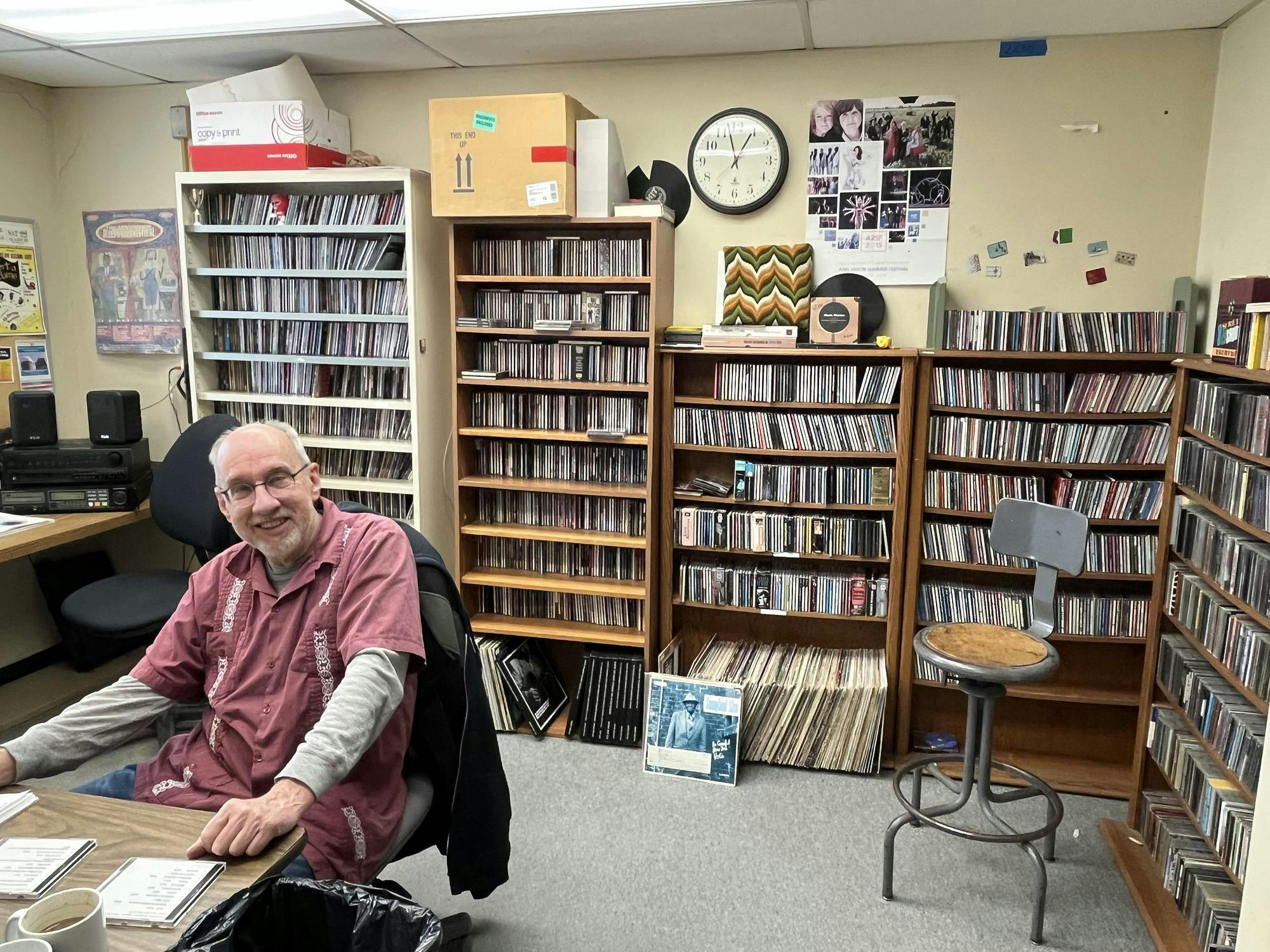A smiling man wearing a red short-sleeved shirt over a long-sleeved gray shirt sits at a brown desk. Behind him are several bookcases filled with CDs and records.