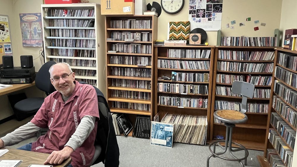 A smiling man wearing a red short-sleeved shirt over a long-sleeved gray shirt sits at a brown desk. Behind him are several bookcases filled with CDs and records.