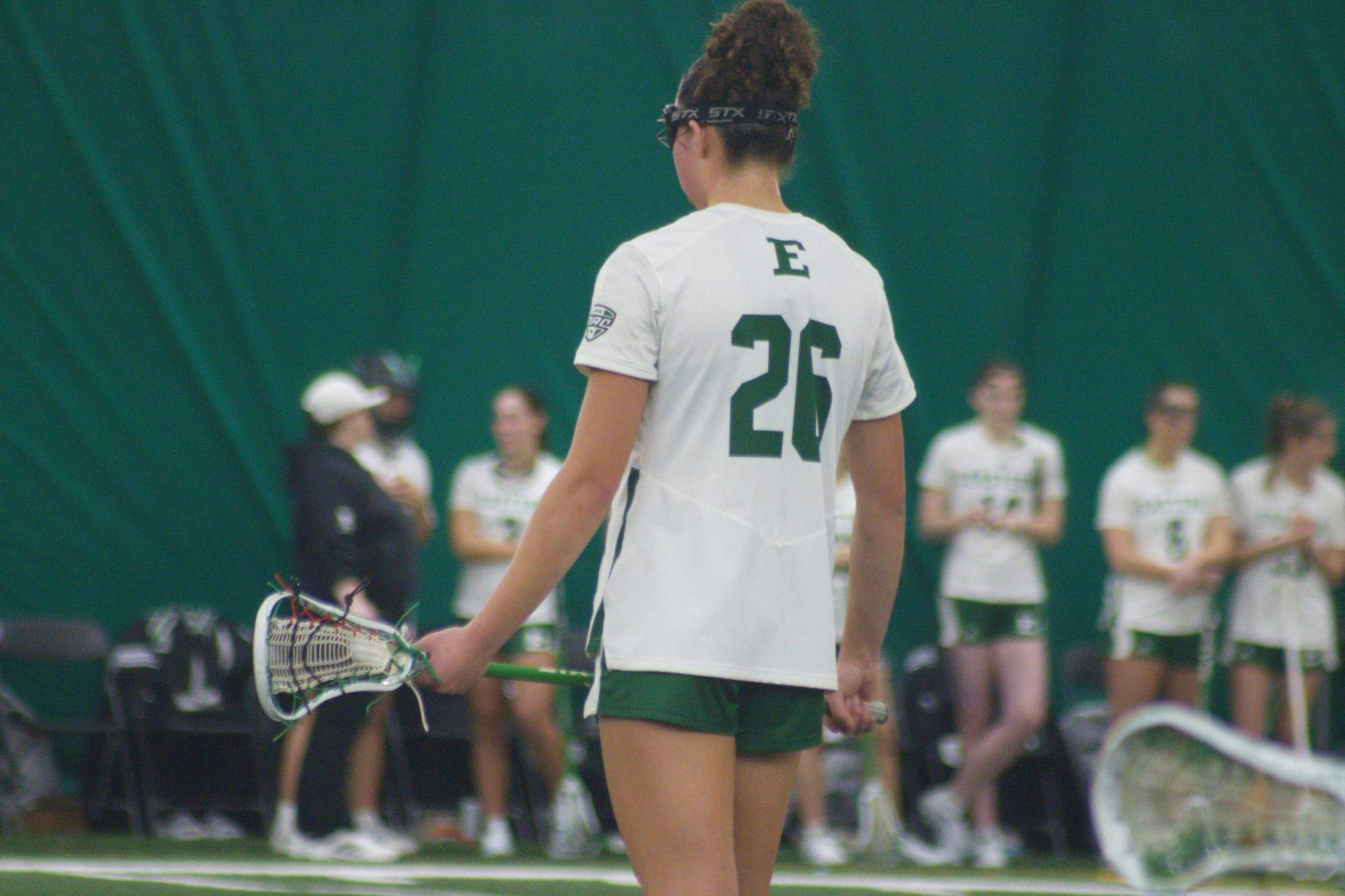 A lacrosse player in a white jersey stands facing away with a large number 26 visible in green text.
