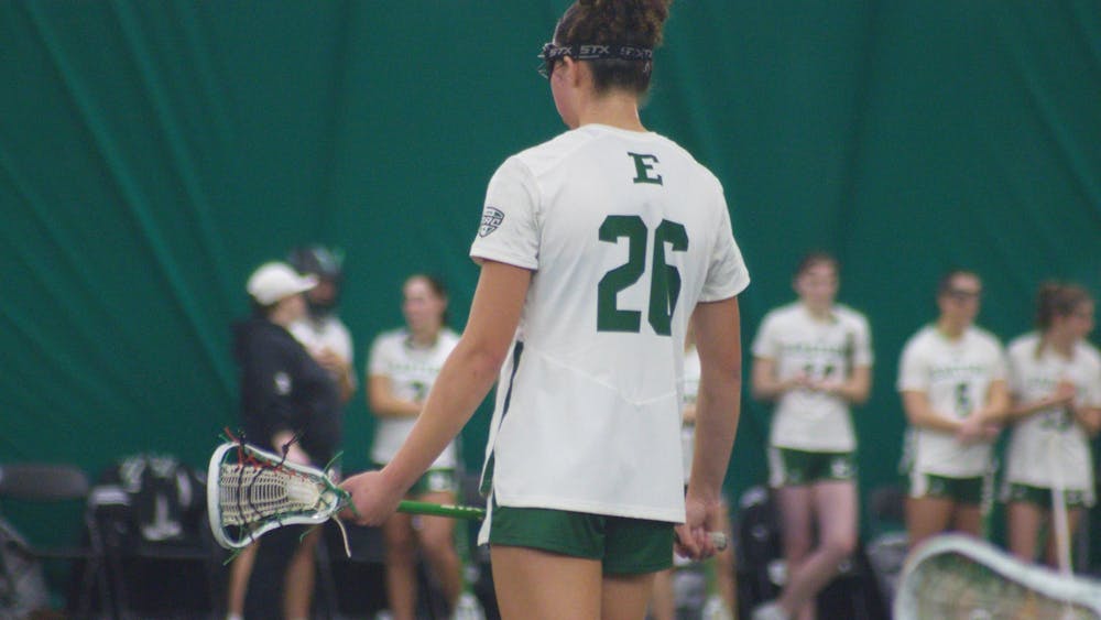 A lacrosse player in a white jersey stands facing away with a large number 26 visible in green text.