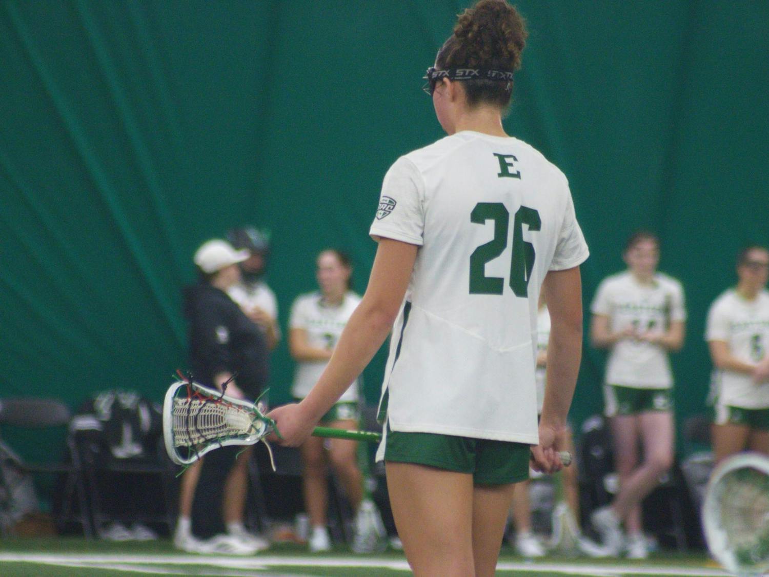 A lacrosse player in a white jersey stands facing away with a large number 26 visible in green text.