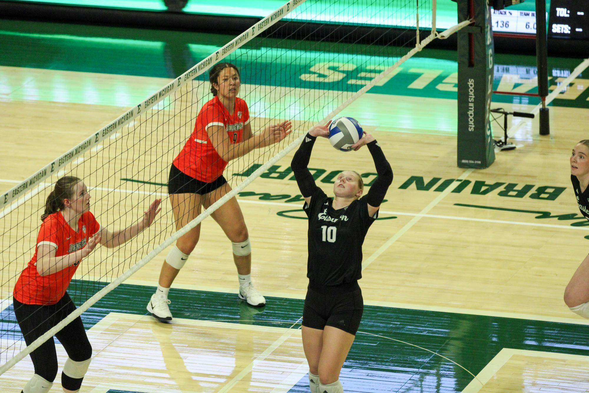#10 Ashlynn Belcher, in a black jersey, setting the volleyball while two BGSU players, in orange jerseys, watch from behind the net. 