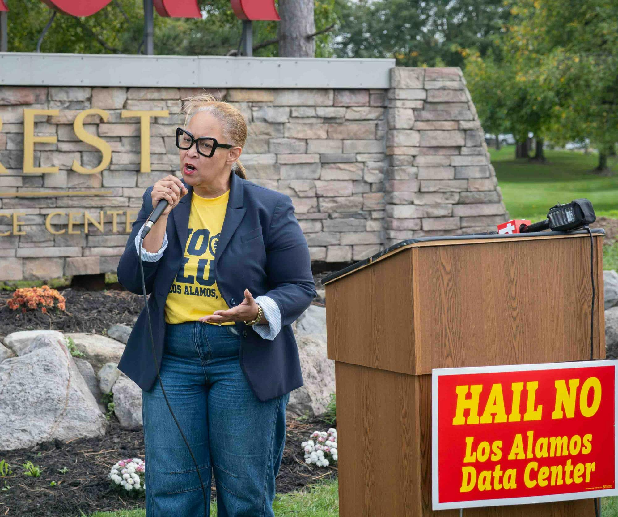  Leah Mills-Chapman stands at a podium outside holding a microphone and wearing a "No Blue and No Los Alamos, Too" T-shirt.