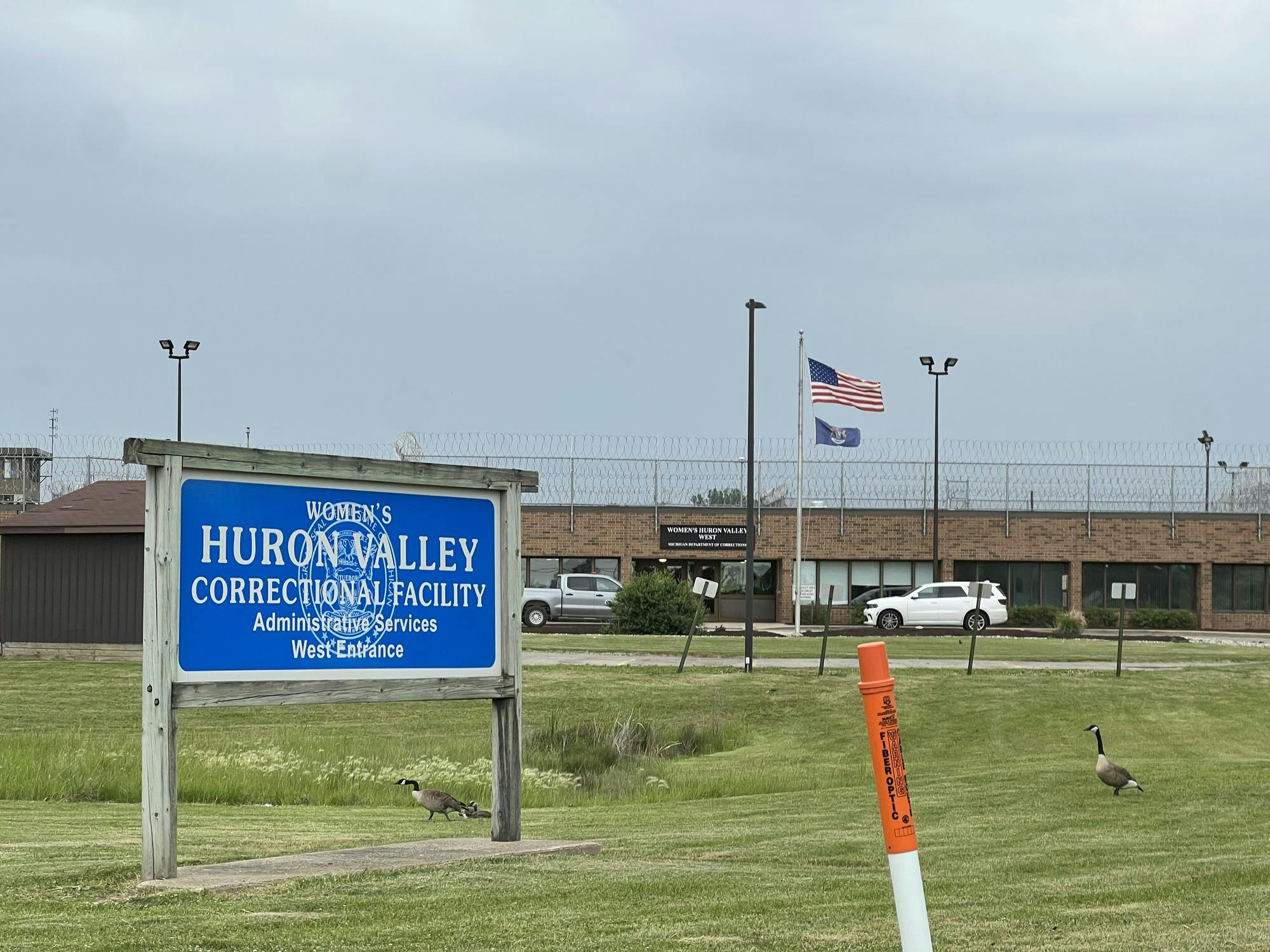 A blue sign with the Michigan seal marks the west entrance of the Women's Huron Valley Correctional facility, a brick building topped with a barbed wire fence.