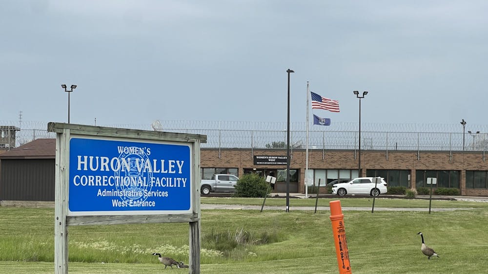 A blue sign with the Michigan seal marks the west entrance of the Women's Huron Valley Correctional facility, a brick building topped with a barbed wire fence.