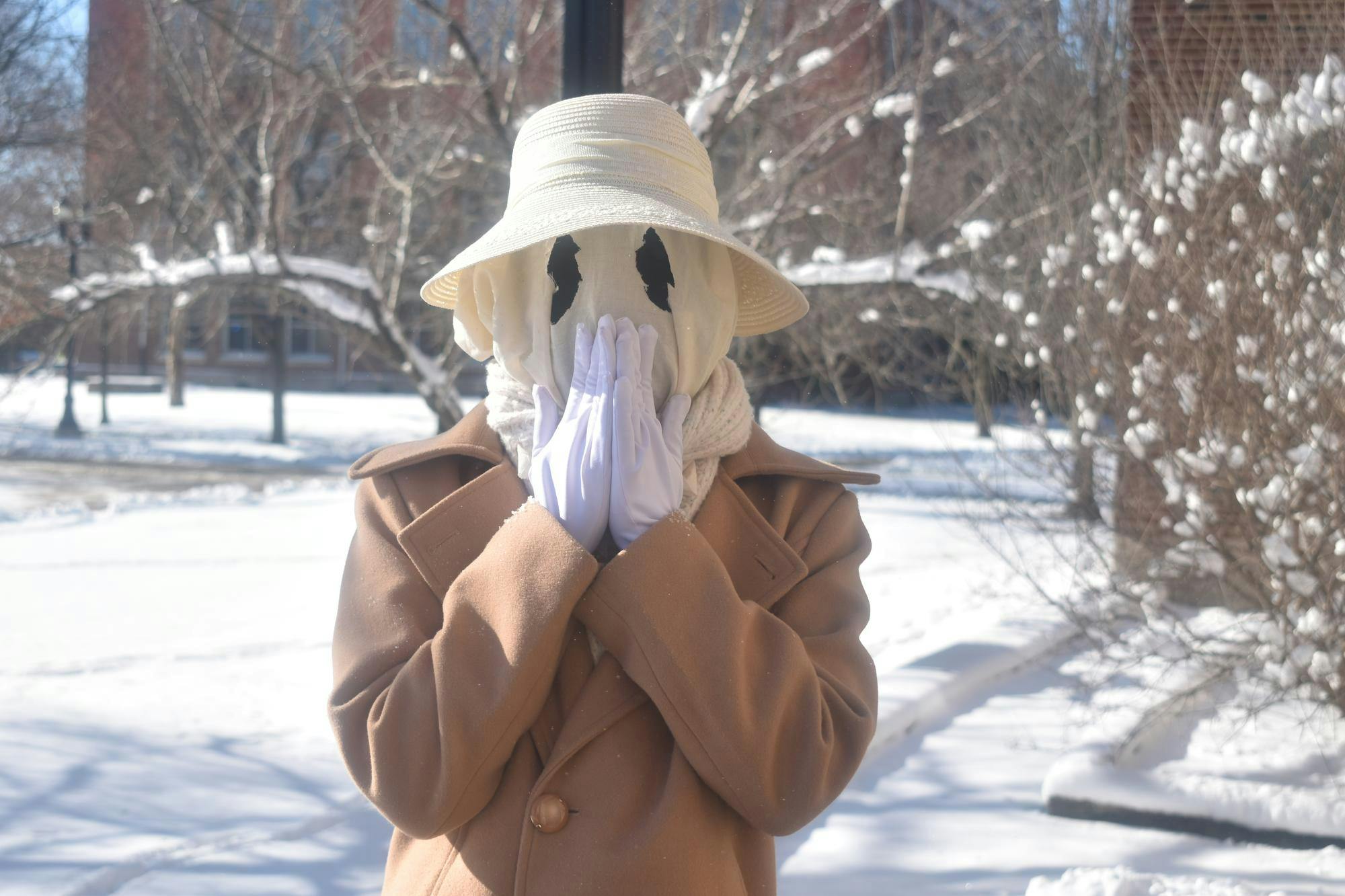 Guinnevere has a white ghostly face mask with black circle eyes, with a white brimmed lace hat and a brown coat, with her hands placed over her mouth.