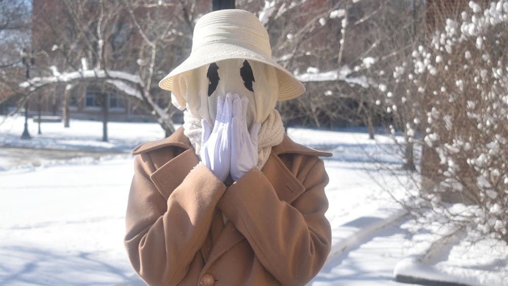 Guinnevere has a white ghostly face mask with black circle eyes, with a white brimmed lace hat and a brown coat, with her hands placed over her mouth.