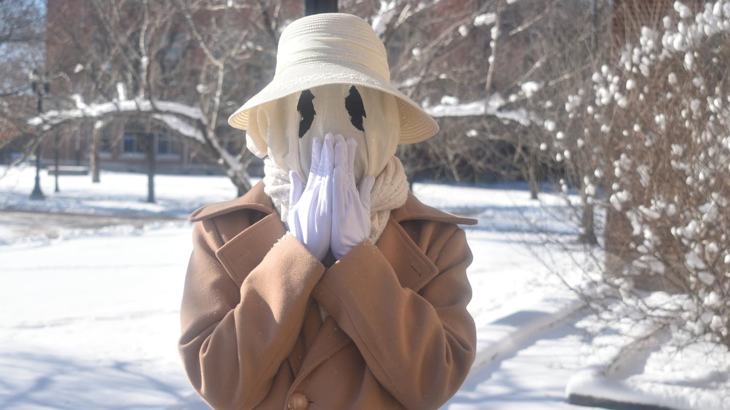 Guinnevere has a white ghostly face mask with black circle eyes, with a white brimmed lace hat and a brown coat, with her hands placed over her mouth.